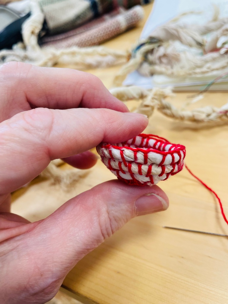 A close up photograph of a hand holding a small vessel shaped object. The object is made of thick white string and has been sewn together with a vibrant red thread. The thread hangs is visible behind the vese=sel, lying on a wooden table, still attached to the needle.