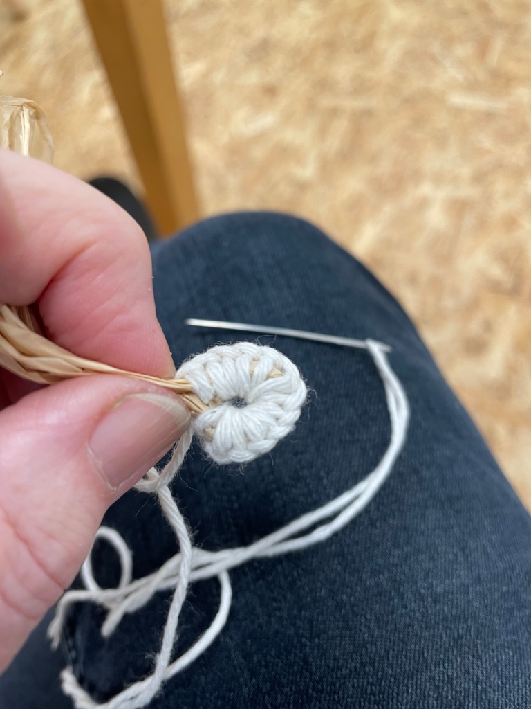 A close up photograph taken from above of needlework held between a finger and thumb and suspended over the persons leg. The needlework has been made with thick white thread and has been created around a loop of natural raffia, making a circular piece of needlework. The thread is still hanging from the needlework and attached to a needle, which rests on the person's leg. The white of the thread stands out against the dark fabric of their jeans.